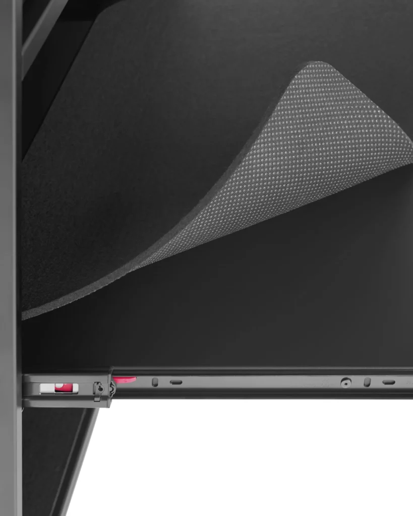 Close-up of a pull-out shelf mechanism inside a metal cabinet, showing a sliding rail with a small red release tab and a textured black liner being lifted to reveal its underside