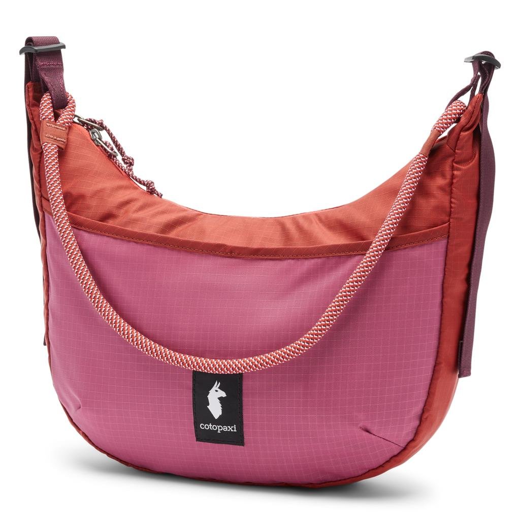 Cotopaxi shoulder bag in rose pink and deep coral ripstop fabric, featuring a curved shape, black front logo patch, and a braided rope strap layered over an adjustable webbing strap
