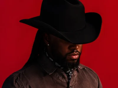 A moody, studio-lit portrait of Shaboozey in profile, partially obscured by shadow, wearing a wide-brimmed black cowboy hat and a dark outfit layered with a subtle chain necklace. The vivid red background creates a striking contrast, casting the figure into a silhouette-like presence while emphasizing the contours of his face and the brim of the hat, evoking a cinematic, Western-inspired aesthetic with a modern, stylized edge