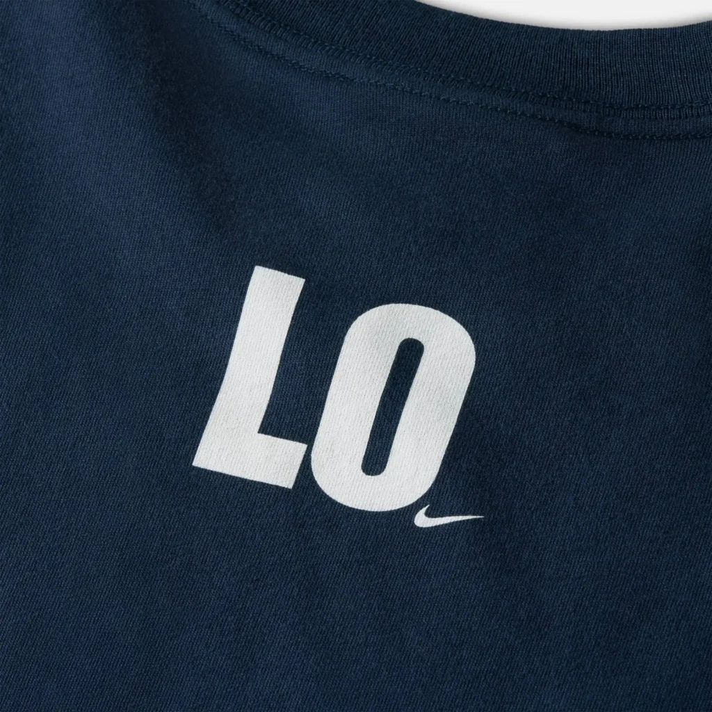Close-up of a navy cotton T-shirt highlighting a crisp white “LO” graphic printed on the chest, accompanied by a small Nike Swoosh beneath. The image emphasizes the smooth fabric texture, clean stitching along the collar, and the minimalist, graphic-led branding