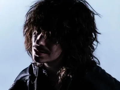 Portrait of Gunnr in low-key studio lighting, with long, tousled dark hair partially covering his face. He faces slightly downward and to the side, wearing a black leather jacket against a soft gray background, creating a moody, introspective atmosphere