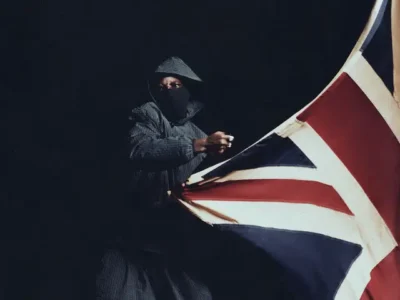 JD Cliffe wears a maskin a dark setting holding and pulling a large Union Jack flag, dressed in textured outerwear with only eyes visible; stark lighting emphasizes contrast, with a “Parental Advisory” label in the corner