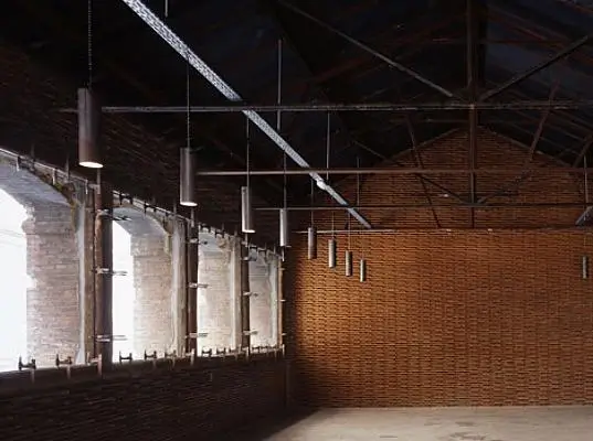 A spacious industrial interior with exposed brick walls and a high, open ceiling is lit by a row of tall arched windows along one side. Suspended cylindrical lights hang in a linear arrangement from the ceiling beams, while natural light streams through the windows, casting soft illumination across the empty floor and emphasizing the warm texture of the brickwork