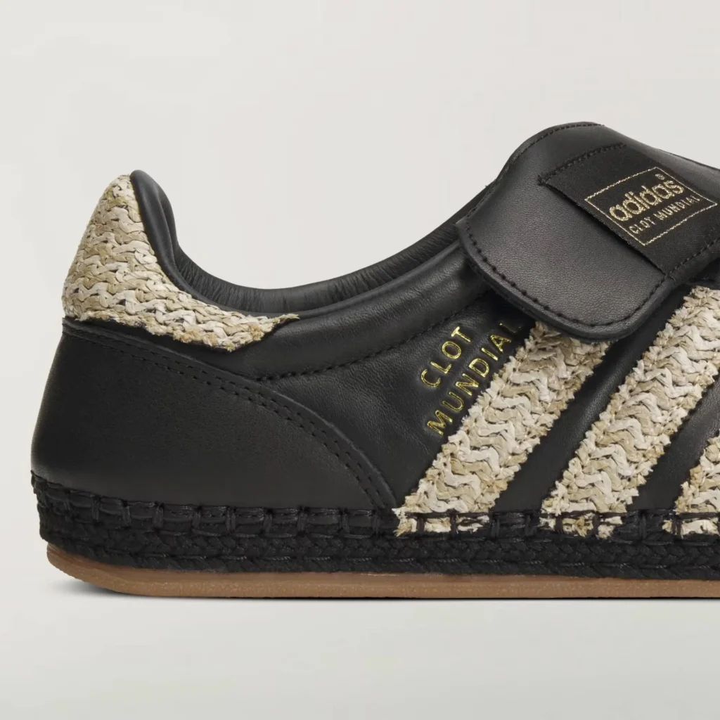 side detail of black adidas x CLOT Mundial sneaker highlighting raffia-woven Three Stripes and heel panel, gold “CLOT Mundial” branding, premium leather upper, and espadrille-style stitched midsole