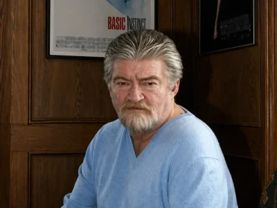 Joe Eszterhas seated in a wood-paneled room, wearing a light blue sweater with a serious expression, framed by Basic Instinct and Showgirls posters behind him in a composed, studio-lit portrait setting