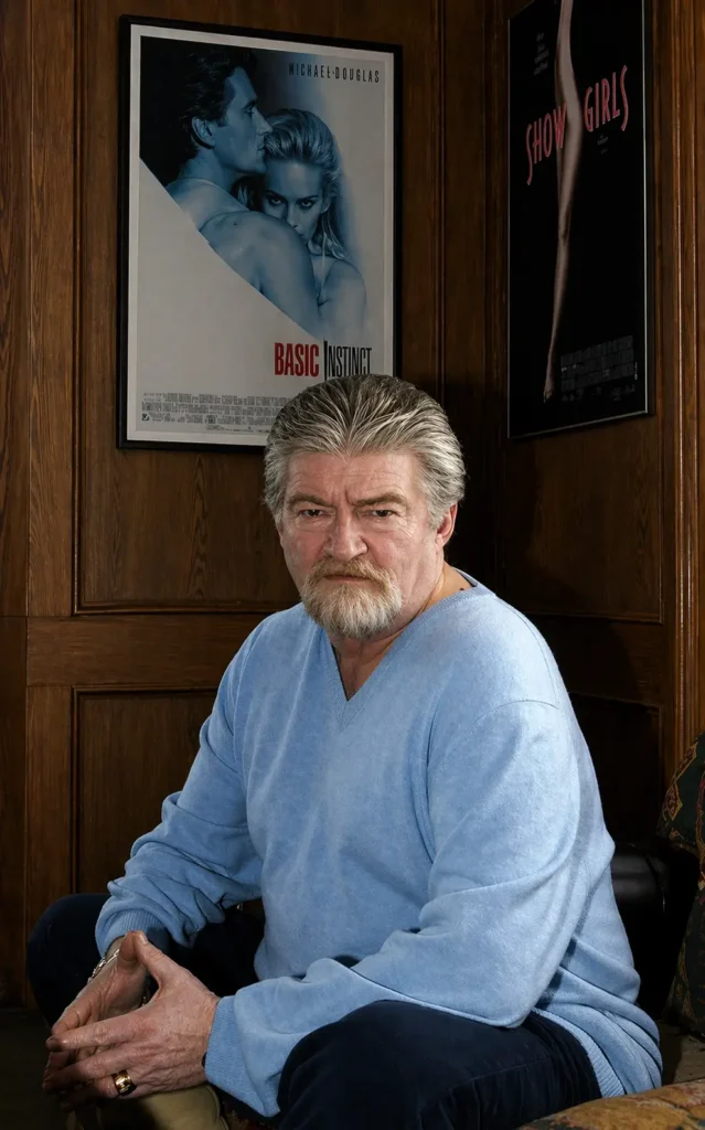 Joe Eszterhas seated in a wood-paneled room, wearing a light blue sweater with a serious expression, framed by Basic Instinct and Showgirls posters behind him in a composed, studio-lit portrait setting