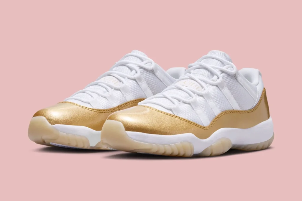 Angled front view of a pair of white low-top sneakers with mesh uppers and metallic gold patent leather mudguards, featuring white laces, cushioned midsoles, and translucent gum outsoles, set against a soft pink background