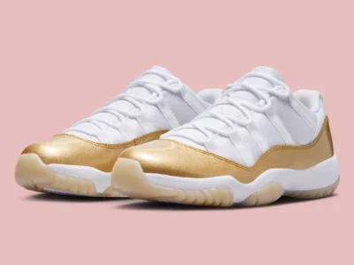 Angled front view of a pair of white low-top sneakers with mesh uppers and metallic gold patent leather mudguards, featuring white laces, cushioned midsoles, and translucent gum outsoles, set against a soft pink background