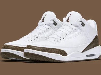 White leather low-top shoe with a clean, tumbled texture upper and perforated detailing across the toe box and mid-panel. The design features tonal white laces and eyelets, paired with a slightly padded collar for structure. A signature brown elephant-print mudguard wraps around the toe and heel, matched with a deep mocha accent on the heel tab and outsole. The midsole remains crisp white with a visible air unit at the heel, while the tongue is finished with a contrasting embroidered logo. Overall, the shoe balances minimal white composition with earthy brown contrasts for a refined, classic look