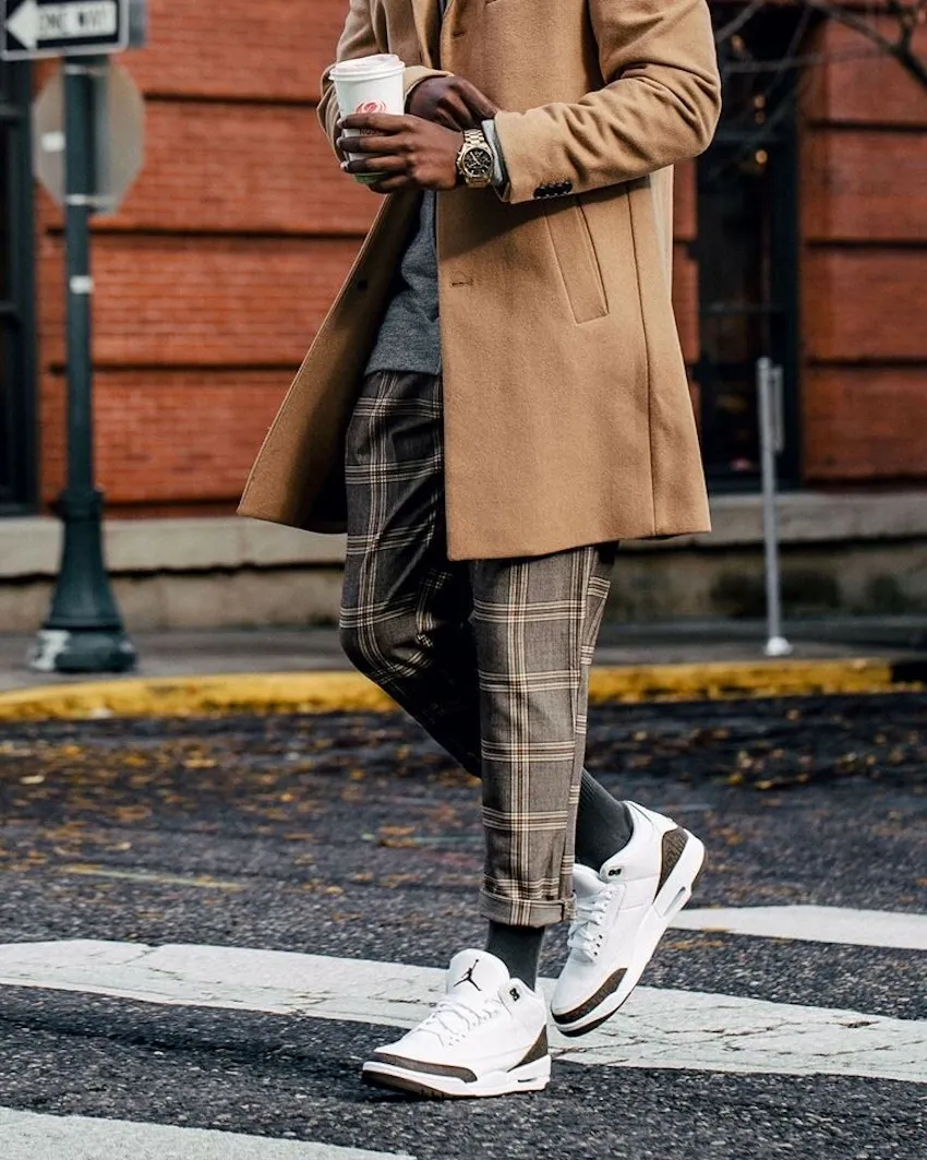 Lifestyle shot of a person walking across a city street wearing white leather sneakers with brown elephant-print accents. The shoes are paired with cropped plaid trousers in muted brown and grey tones, allowing the sneakers to remain visible and prominent. A camel overcoat drapes over a layered outfit, complemented by grey socks and a casual knit underneath. The wearer holds a takeaway coffee cup, adding to the relaxed, urban feel. The sneakers’ clean white upper contrasts with the darker street surface, while the mocha-toned detailing ties seamlessly into the earthy palette of the outfit. The scene is set against a brick building backdrop, reinforcing a polished, everyday street-style aesthetic