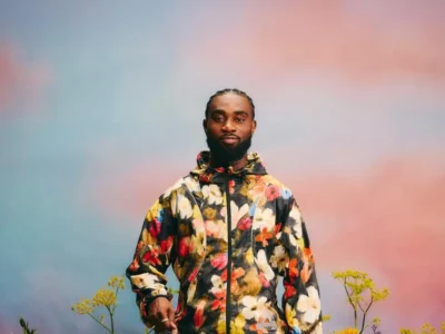Kofi Owusu-Ansah stands centered against a soft gradient sky of pastel blue and warm pink, wearing a floral-patterned jacket that echoes natural motifs, his calm expression reinforcing the reflective, grounded tone associated with “Gardener Flow”