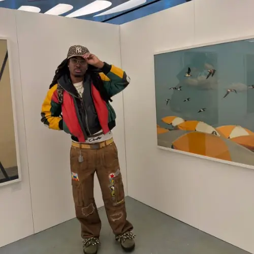 Cochise stands in a gallery space wearing a multicolored jacket, brown patchwork pants, and a patterned cap, posing beside a framed artwork of birds flying over orange-toned forms against a room background