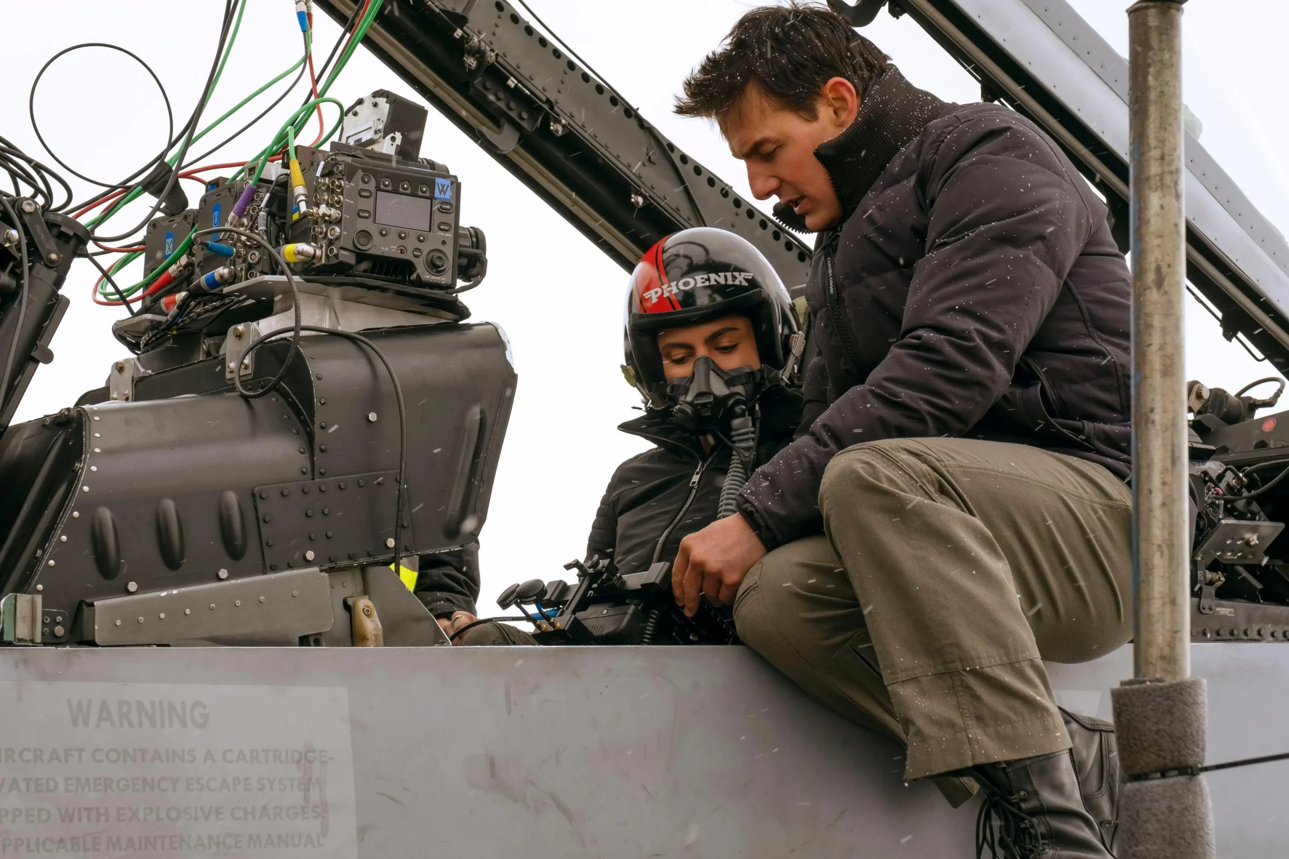 Tom Cruise seated on the edge of a fighter jet cockpit during filming, adjusting equipment beside a helmeted co-pilot, with visible camera rigging and wiring highlighting the practical, in-air production setup of Top Gun: Maverick