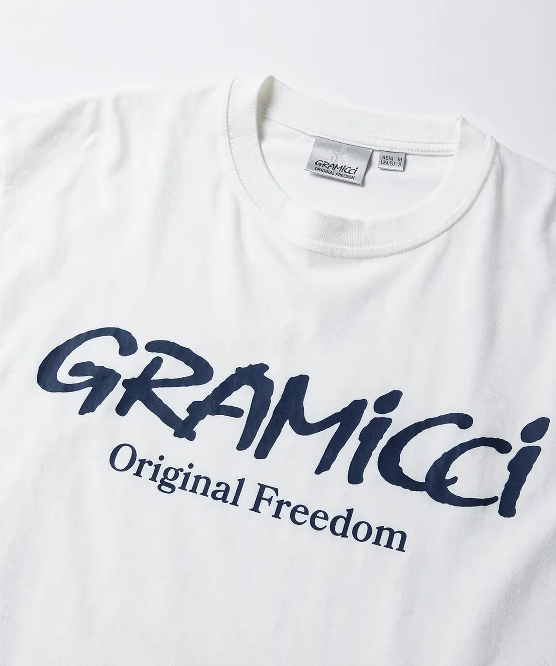 High-resolution flat lay of a white Gramicci T-shirt with a ribbed crewneck, featuring a bold navy script logo and “Original Freedom” text on a clean studio background