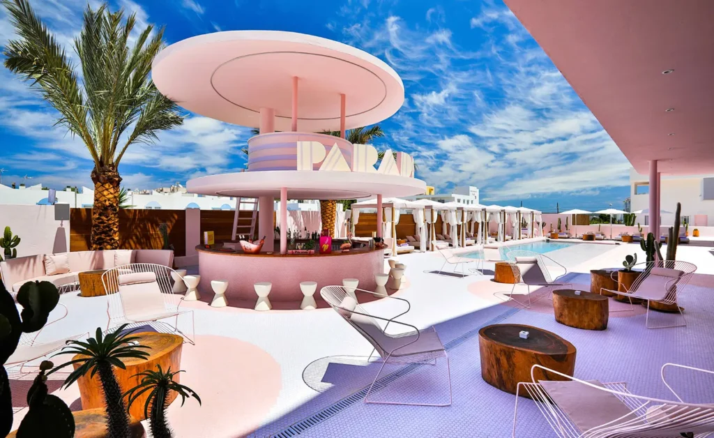 Pastel pink poolside bar pavilion at Los Felices Ibiza featuring a circular canopy structure, lounge seating, palm trees, and cabana-lined pool area under a vivid blue sky