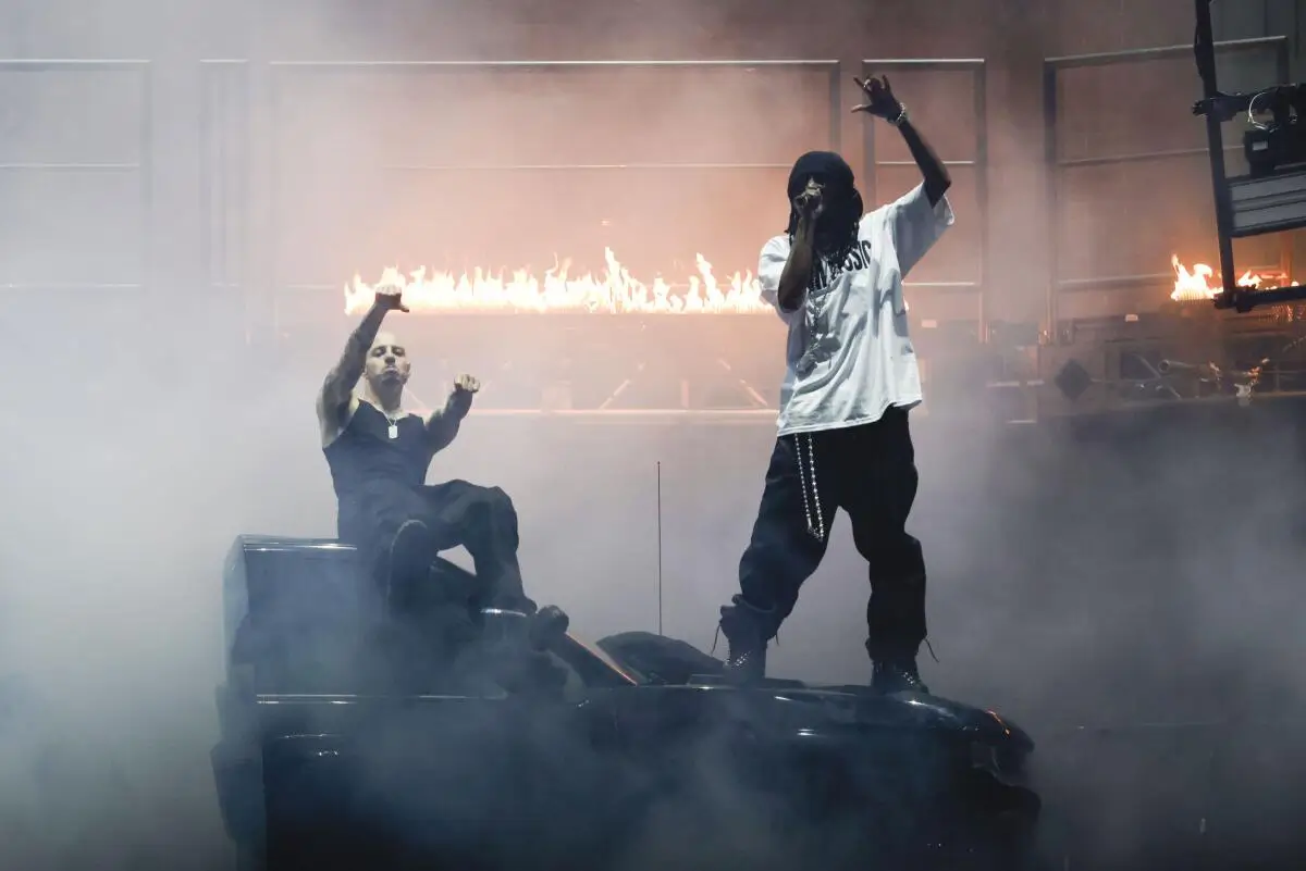 Two performers on stage amid heavy smoke and a backdrop of flames, one standing and rapping into a microphone while the other sits on a vehicle raising a fist, both framed by intense lighting and pyrotechnics