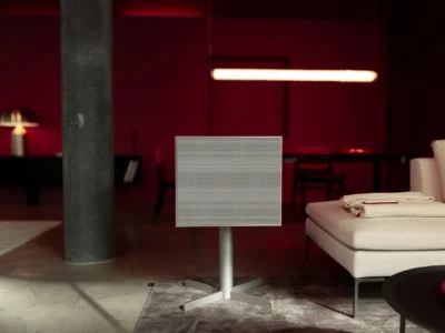 Geneva Lab DeCon/XL speaker in a clean white finish on a minimalist stand, centered in a moody modern interior with deep red ambient lighting, a lounge sofa, and sculptural furnishings creating a cinematic, design-forward atmosphere