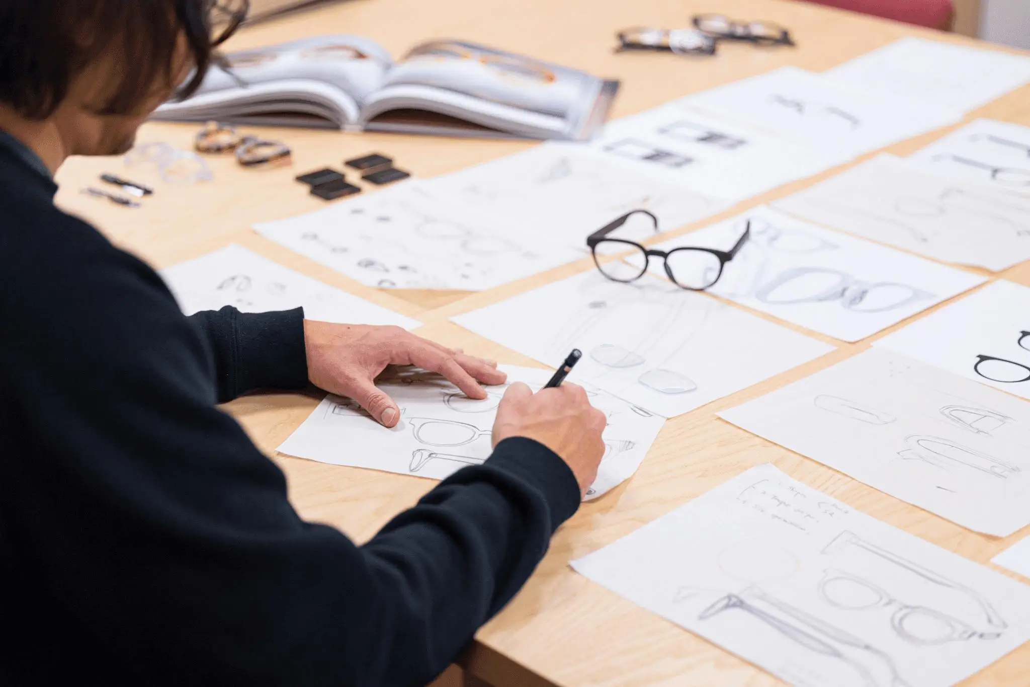 Designer seated at a wooden table sketching eyeglass concepts on paper, surrounded by multiple layout drawings, a prototype pair of glasses, small material samples, and an open reference book, captured in a focused studio workspace