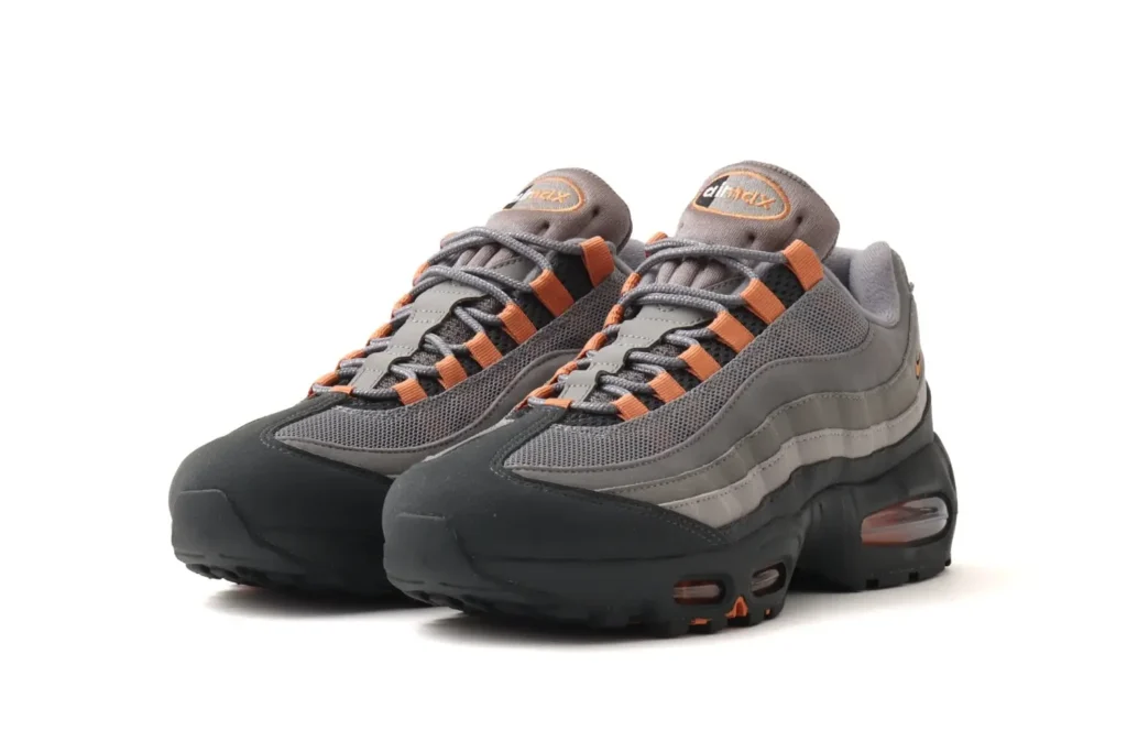 A pair of Nike Air Max 95 shoe in muted grey tones with black mudguards and subtle orange accents, featuring layered mesh and suede panels, rope-style laces with orange eyelets, and a visible Air unit embedded in the dark outsole, presented against a clean white background