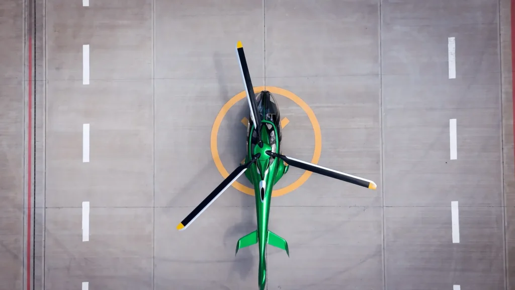 Top-down view of a sleek green helicopter positioned on a circular helipad, its rotor blades extended symmetrically against a clean concrete surface marked with lane-style lines