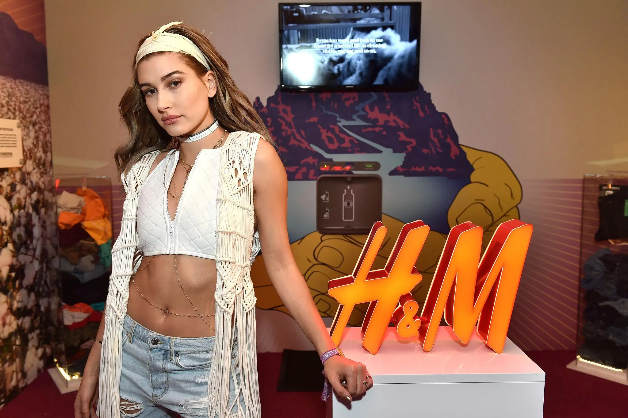 Hailey Bieber poses at an H&M brand activation event, wearing a stylish boho-chic outfit featuring a white macramé fringe vest, quilted crop top, and light-wash ripped jeans. She stands confidently beside a large illuminated orange H&M logo, with a screen and desert-themed backdrop behind her