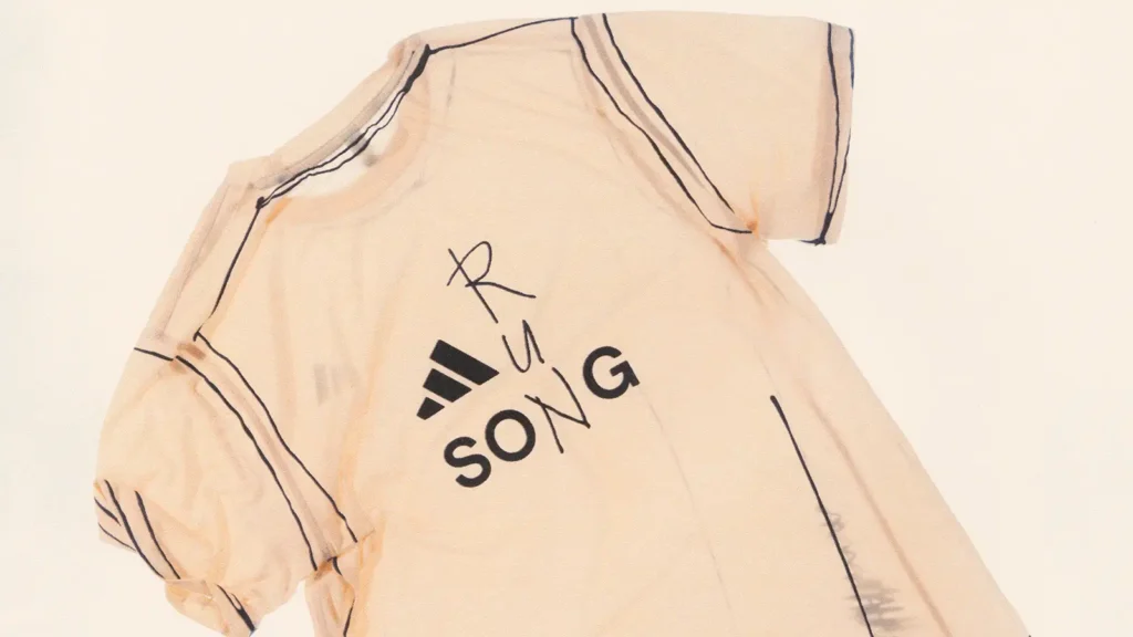 Close-up of Song for the Mute x adidas Running SS26 technical tee in a soft beige tone, featuring co-branded adidas and “SONG” logo with hand-drawn style graphic detailing and visible seam lines