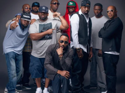 Wu-Tang Clan group portrait featuring all core members standing and crouching together against a neutral studio backdrop, wearing casual streetwear including caps, hoodies, and sunglasses, with RZA positioned at the front in a crouched pose, surrounded by Method Man, Raekwon, Ghostface Killah, Inspectah Deck, GZA, U-God, Masta Killa, and Cappadonna, conveying a unified yet individual presence through varied stances and expressions