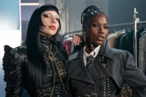 Lady Gaga and Doechii pose backstage in a fashion setting, styled in structured black looks—Gaga in a gothic, high-neck leather ensemble with blunt bangs, Doechii in a tailored suit with braided detailing—standing close with a composed, high-fashion gaze
