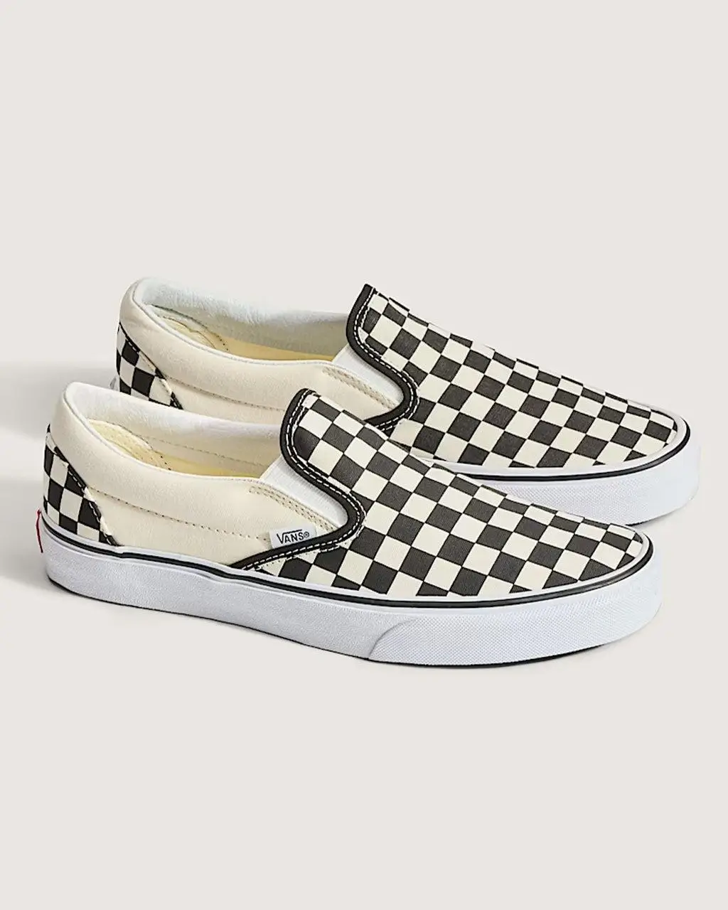 A pair of classic Vans slip-on sneakers in a black-and-white checkerboard pattern, shown in a clean studio setting. The canvas uppers feature the iconic alternating square print, complemented by off-white side panels and elastic gores for easy wear. The shoes sit on a white rubber sole with a textured foxing and a thin black stripe, while a small Vans tag is visible along the side seam