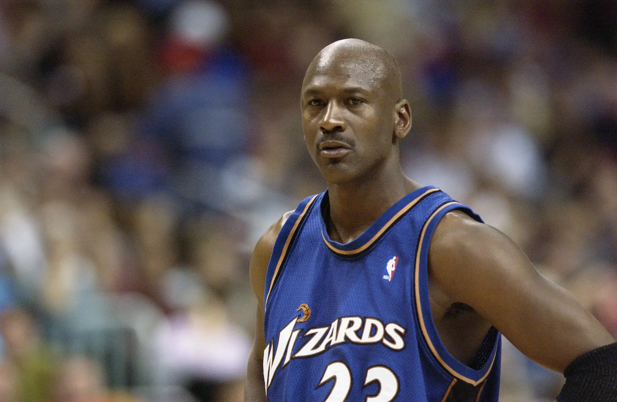 Close-up of Michael Jordan wearing a blue Washington Wizards jersey (#23), standing on the court with a focused expression, sweat visible on his face, and a blurred arena crowd in the background