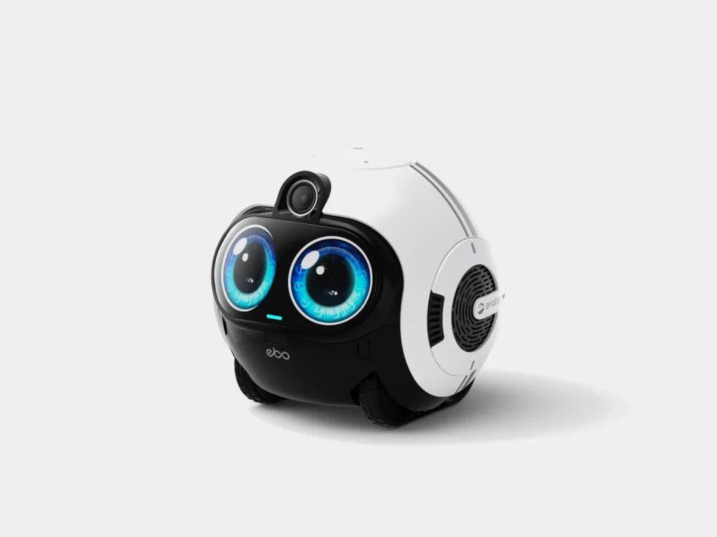A compact, rounded companion robot with a glossy black-and-white body sits on small wheels, featuring oversized animated blue eyes on a digital display, a small front camera perched above like a head, and side speaker grilles—designed with a playful, pet-like aesthetic against a clean white background