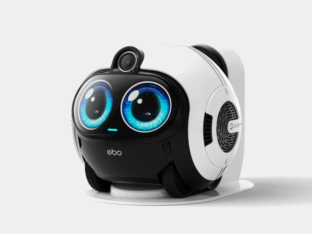 A small, wheeled companion robot with a glossy black front and white shell rests on its charging dock, featuring large expressive blue digital eyes, a raised camera module like a head, and side speaker vents—designed with a friendly, pet-like appearance in a clean studio setting