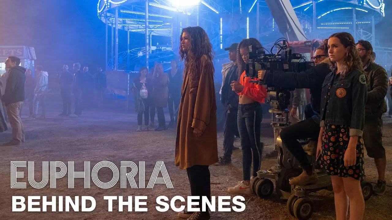 Behind-the-scenes moment on a film set at a nighttime carnival, with bright neon rides glowing in the background. A camera operator films a young woman Zendaya standing in a long brown coat while crew members and extras gather around. Another young woman stands in the foreground watching, as large on-screen text reads “Euphoria Behind the Scenes,” emphasizing the production setting and atmosphere