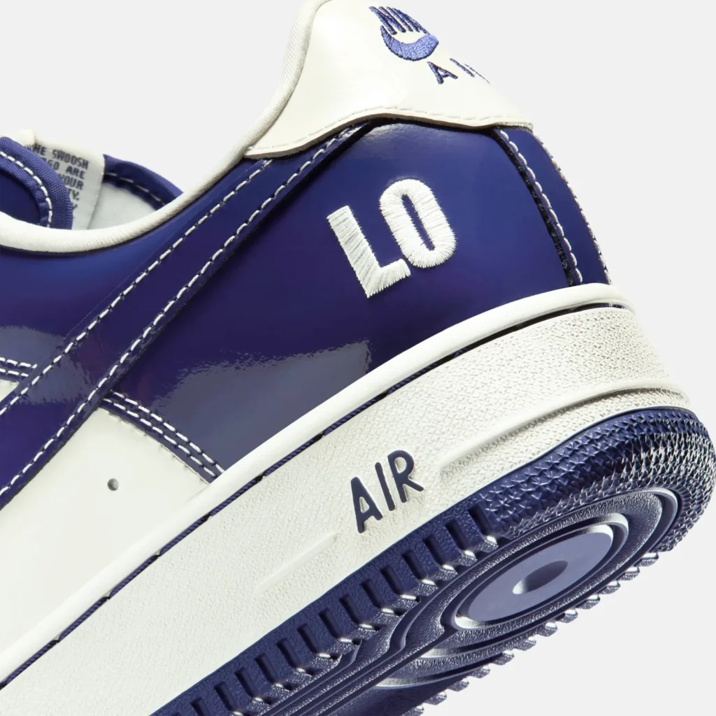 Close-up detail of a Nike Air Force 1 shoe in a glossy deep blue and white colorway, highlighting the heel area with embroidered “LO” lettering. The image emphasizes contrast stitching, textured white midsole with “AIR” branding, and the ridged blue outsole, showcasing premium material finish and precise construction