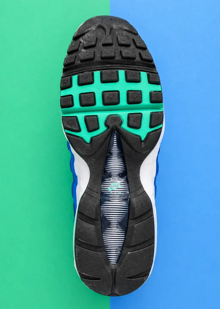 Bottom view of a Nike Air Max 95 outsole with black rubber tread and teal grid detailing, centered on a split green and blue surface, showing traction patterns and midfoot branding in a crisp show