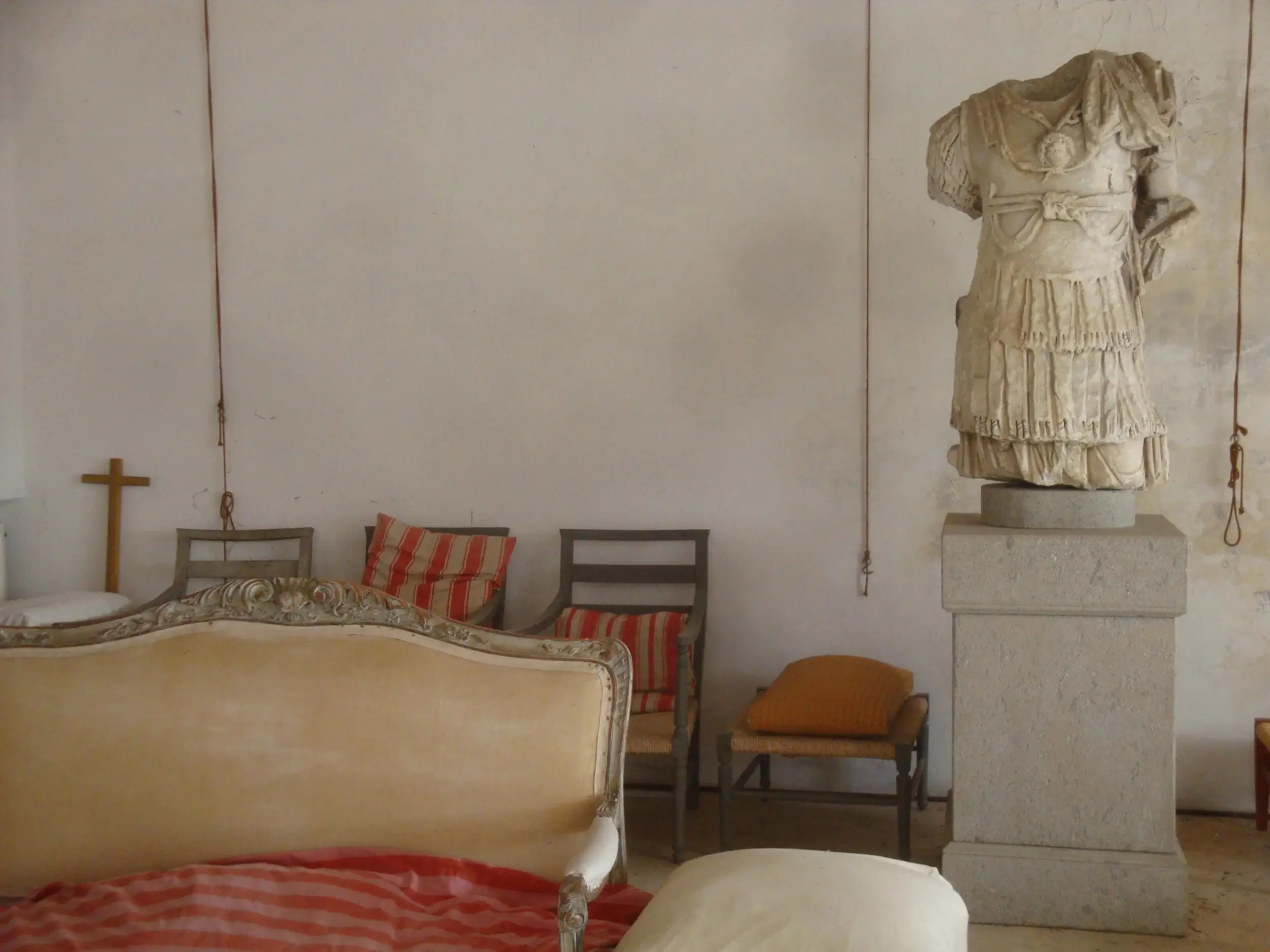 An understated, rustic room features a cream-upholstered antique sofa and simple wooden chairs with striped cushions, while a headless classical statue torso stands elevated on a stone pedestal; a small cross on the wall and worn plaster surfaces add to the historical, contemplative setting
