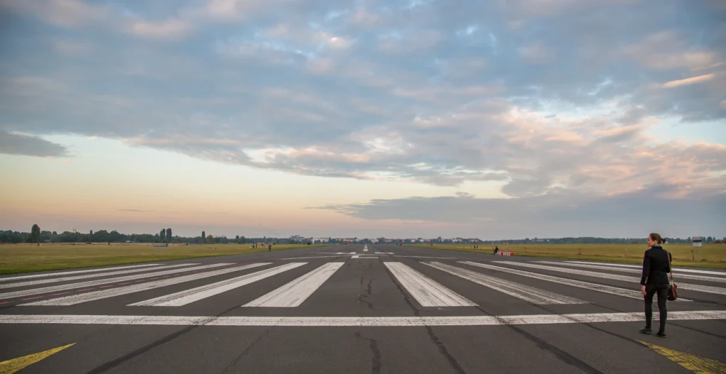 Wide view of an airport runway at sunset with painted markings stretching into the distance under a cloudy sky, with a person walking along the edge