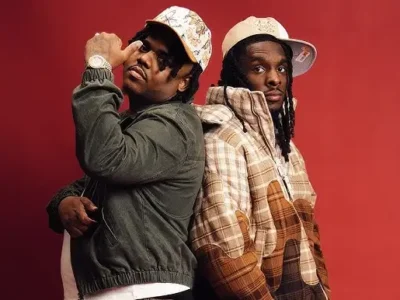 Two men stand back-to-back against a bold red studio background, posing with confident, relaxed expressions. Both wear patterned baseball caps and layered streetwear—one in a dark green jacket with a watch, the other in a patchwork-style plaid jacket in beige and brown tones. Their stance and styling convey a coordinated, fashion-forward streetwear aesthetic with a focus on texture, layering, and attitude
