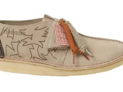 Beige suede Clarks-style Desert Trek shoe featuring abstract line-art graphics across the upper, accented with mismatched laces—one wrapped in orange cord and the other in textured rope—alongside a pink branded tag and a translucent crepe sole