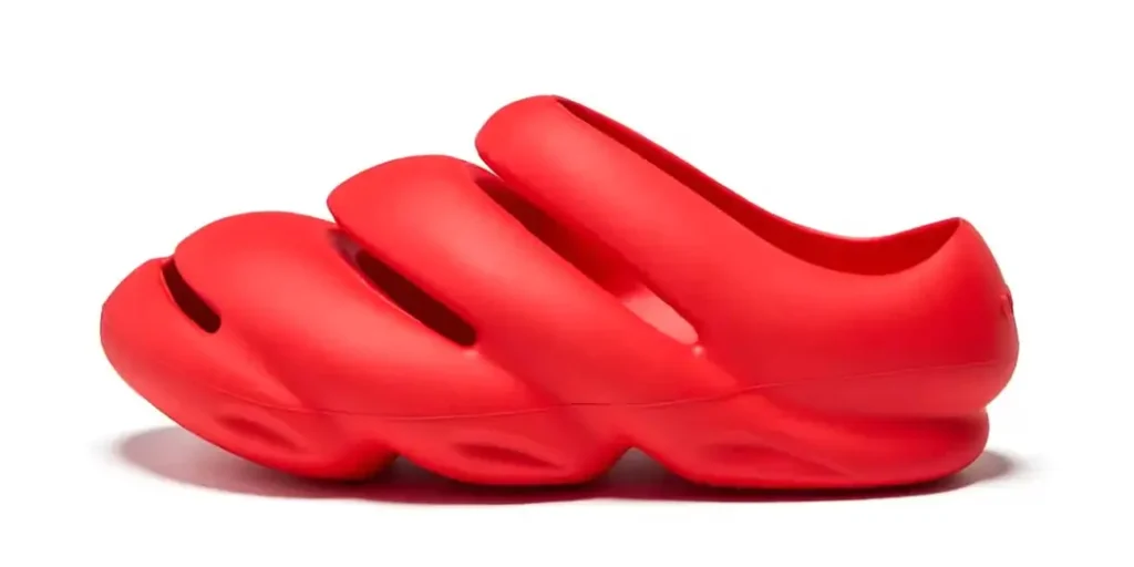 A sculptural, slip-on clog in a vivid monochrome red, featuring a futuristic, organic silhouette with three elongated oval cutouts across the upper. The shoe appears molded from a single piece of foam or rubber, with a smooth matte finish and exaggerated, wave-like contours along the midsole and outsole. Its design leans heavily into biomorphic aesthetics—bulbous, flowing, and almost liquid in form—blurring the line between footwear and industrial design object. The heel is rounded and enclosed, while the forefoot maintains a closed-toe structure, giving it a protective yet avant-garde profile.