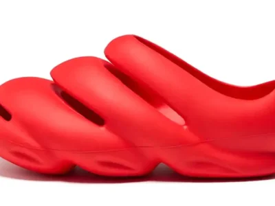 A sculptural, slip-on clog in a vivid monochrome red, featuring a futuristic, organic silhouette with three elongated oval cutouts across the upper. The shoe appears molded from a single piece of foam or rubber, with a smooth matte finish and exaggerated, wave-like contours along the midsole and outsole. Its design leans heavily into biomorphic aesthetics—bulbous, flowing, and almost liquid in form—blurring the line between footwear and industrial design object. The heel is rounded and enclosed, while the forefoot maintains a closed-toe structure, giving it a protective yet avant-garde profile.