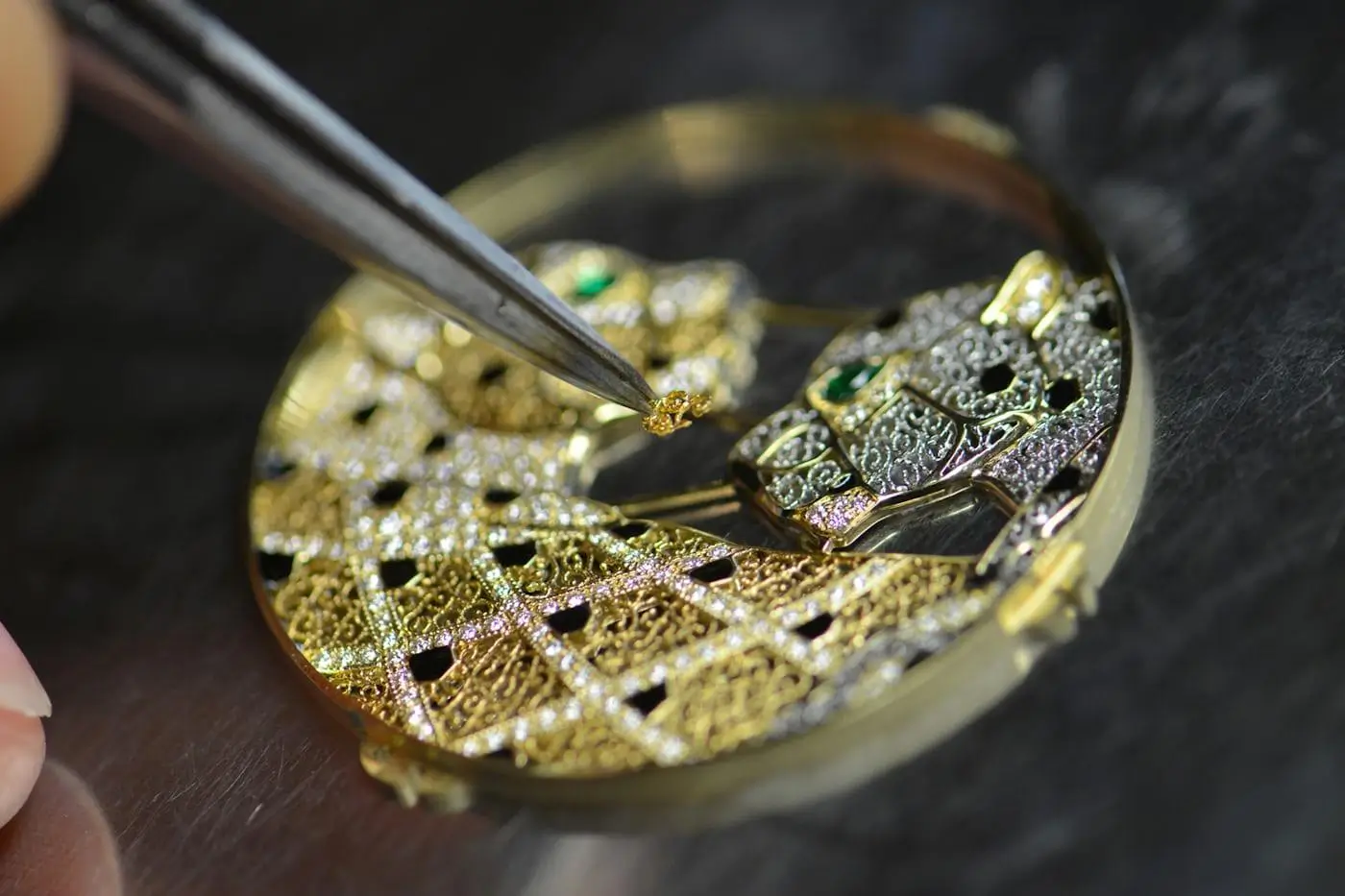 Fine tweezers placing a tiny gold component onto an intricately engraved watch dial, highlighting delicate hand assembly and high-precision decorative craftsmanship