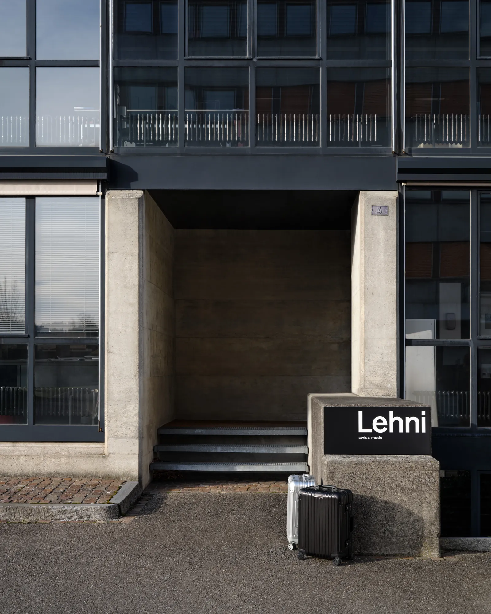 Modern industrial building entrance with concrete pillars and large grid windows, featuring a “Lehni” sign on a low concrete block, with two small hard-shell suitcases placed on the ground nearby