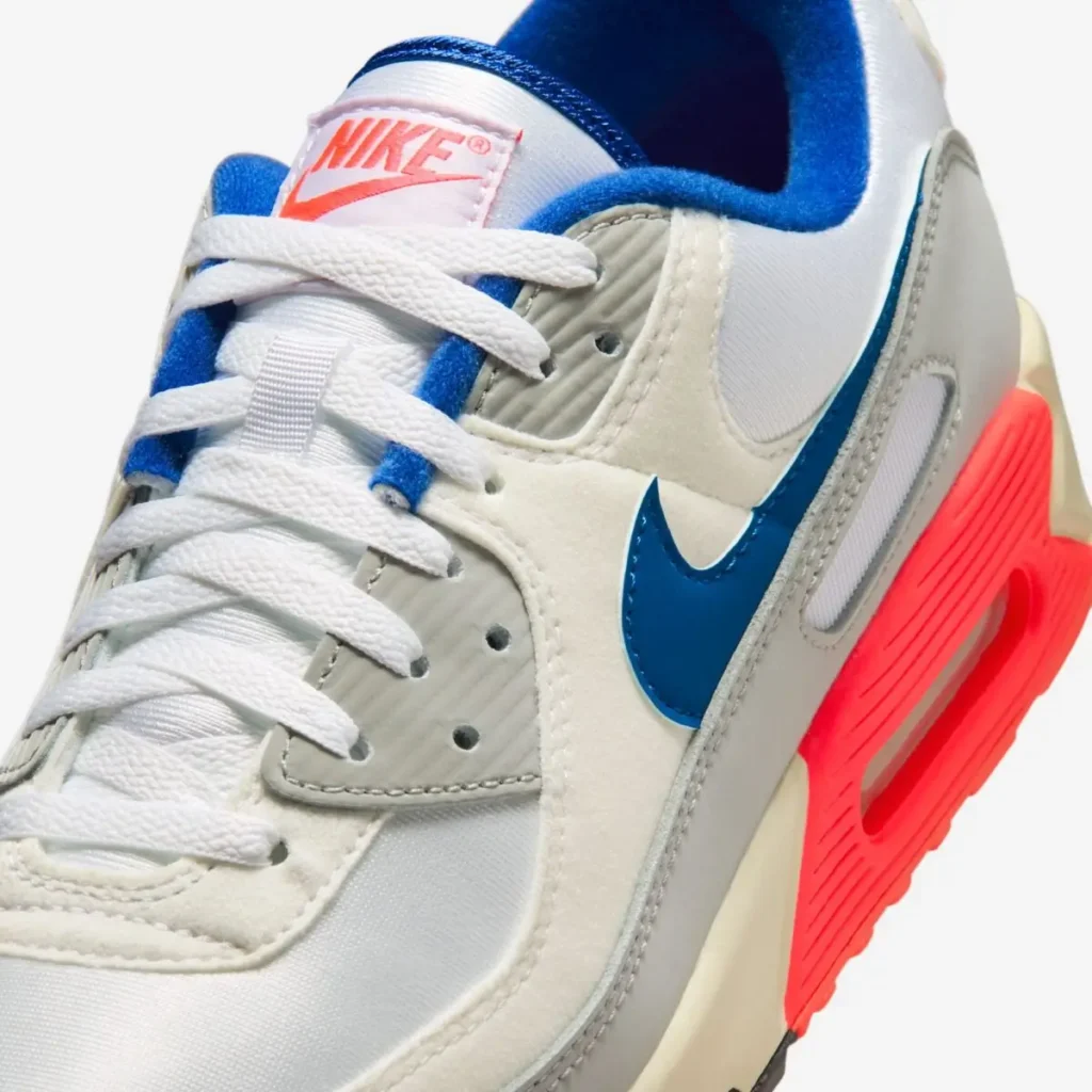 Close-up detail of the Nike Air Max 90 “Ultramarine,” highlighting white laces, textured grey eyelets, blue Swoosh, soft off-white suede overlays, and the vivid red midsole with visible Air cushioning unit