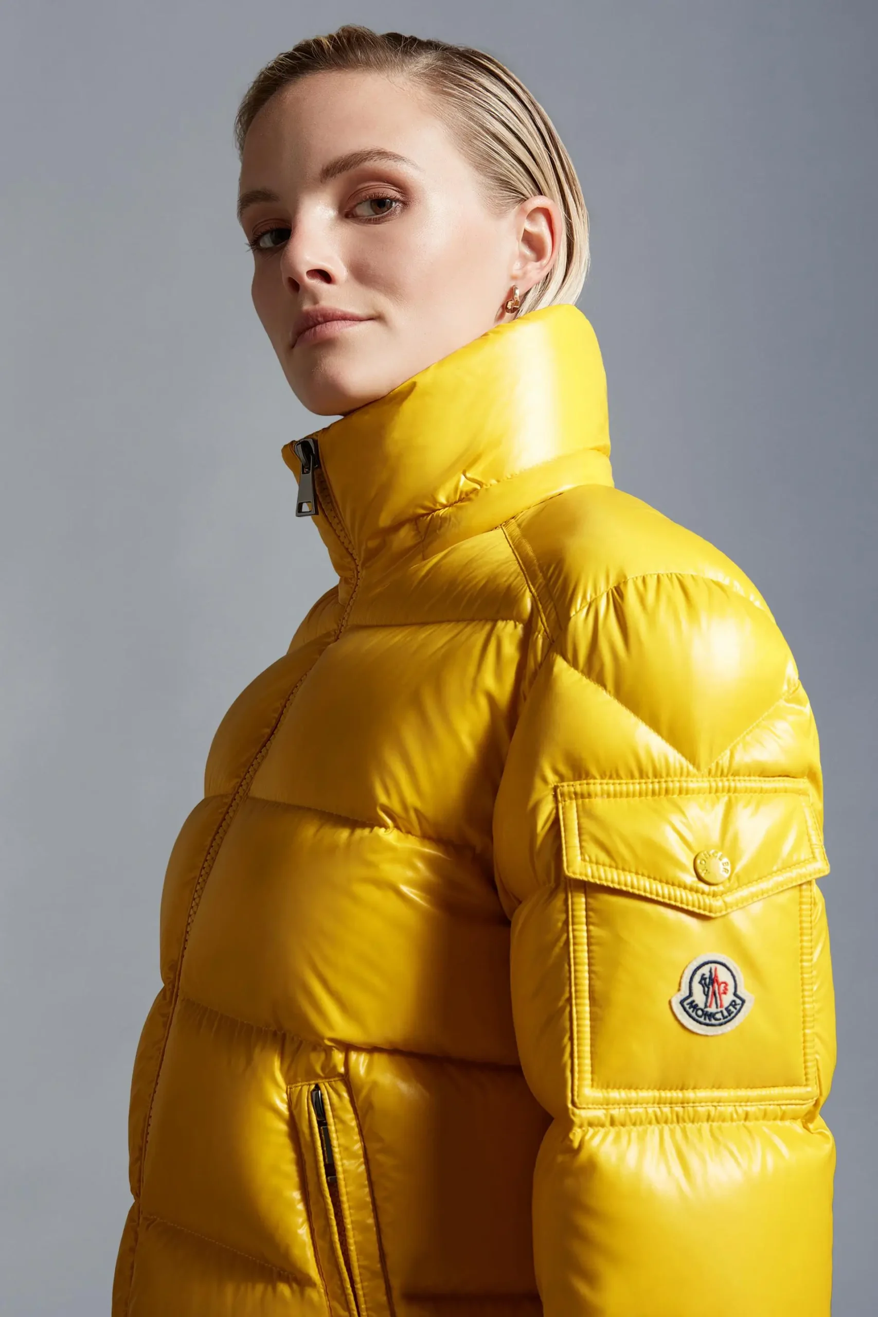 Close-up portrait of a model wearing a glossy sunflower-yellow quilted puffer jacket by Moncler, featuring a high stand collar, structured paneling, and a sleeve utility pocket with the signature logo patch, emphasizing bold color and sculpted volume