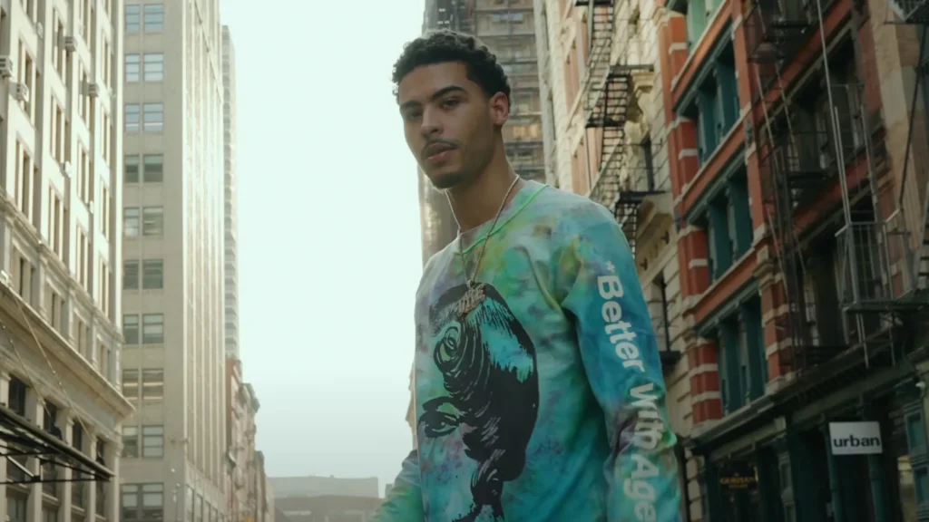 Jay Critch stands on a sunny New York City street lined with tall buildings and fire escapes, looking directly at the camera. He wears a vibrant blue-green tie-dye long sleeve shirt featuring a large black graphic on the chest and white text reading 'Better With Age' on the sleeve