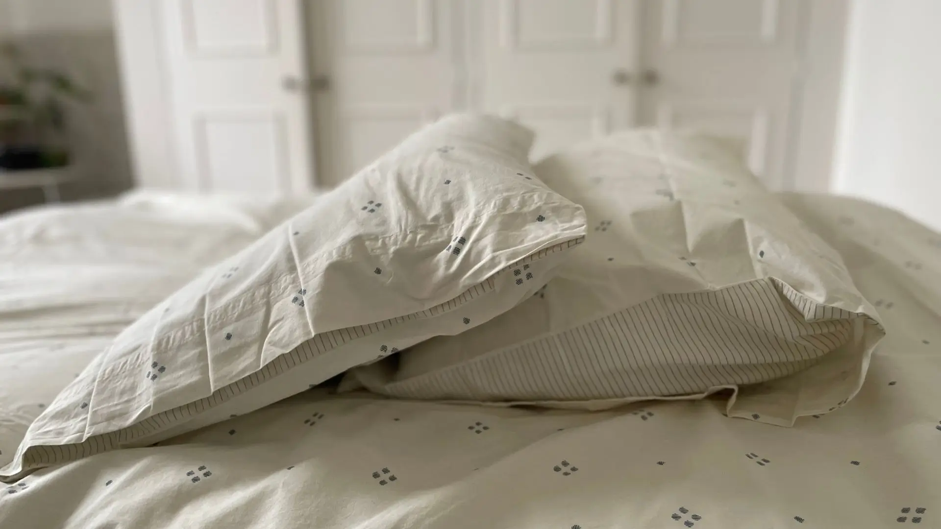 Organic cotton poplin bedding set featuring a subtle rowan berry–inspired dot pattern, with pillowcases showing striped envelope closures, styled on a soft neutral bed in a minimalist bedroom setting