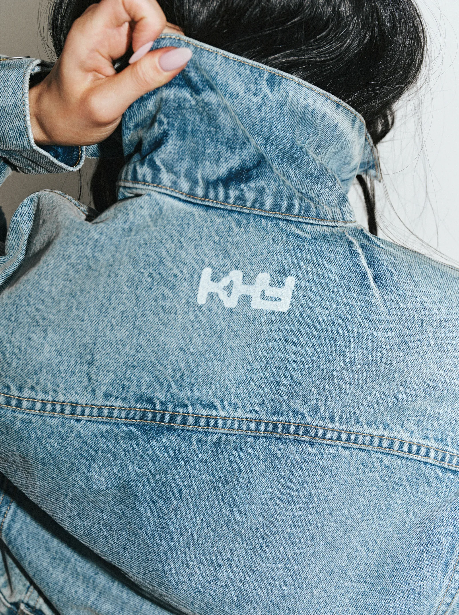 Light-wash denim jacket featuring a white Khy logo embroidered across the upper back, worn by a model lifting the collar, emphasizing texture, stitching, and a relaxed, Los Angeles-inspired aesthetic