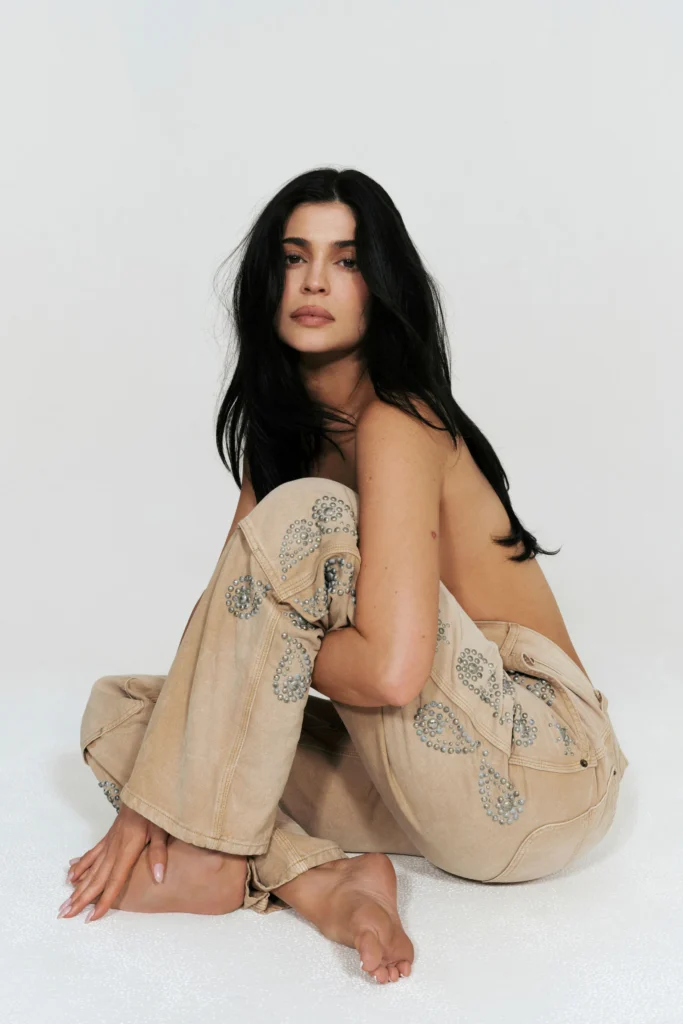 Kylie Jenner poses barefoot in a studio setting for the 'Born in LA' collection by her refreshed fashion brand Khy. She wears a matching beige studded denim jacket and jeans set with intricate silver embellishments, sitting confidently with long dark hair and minimal makeup. Clean editorial campaign shot highlighting the elevated everyday staples from the SS26 refresh, April 2026