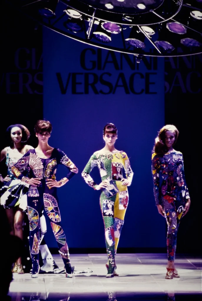 Archival runway show featuring models in bold, graphic Versace designs beneath a “Gianni Versace” backdrop, showcasing the designer’s vibrant, high-impact aesthetic
